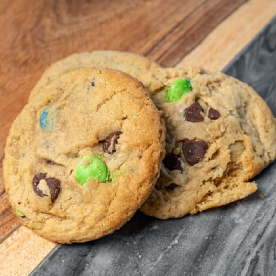 Two CARS Eatery chocolate chip cookies with green and blue candies on a wooden and marble surface.