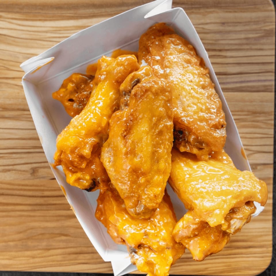 A white takeout container filled with golden-brown, saucy chicken wings on a wooden surface.