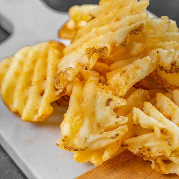 A close-up of golden-brown waffle fries, lightly seasoned, piled on a white and wooden board.
