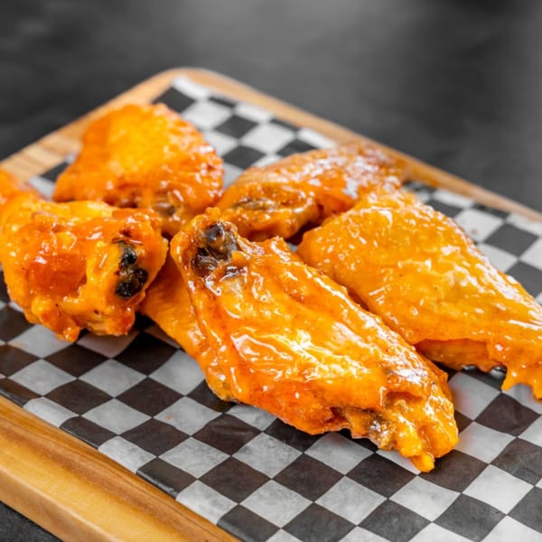 Five buffalo chicken wings, glistening with orange sauce, on checkered paper and a wooden board.