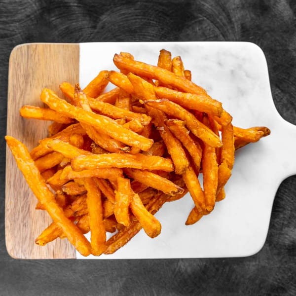 A pile of golden-brown sweet potato fries on a white and wood cutting board.