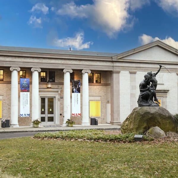 A classical building with columns and banners, a bronze statue on a rock, and a green lawn.