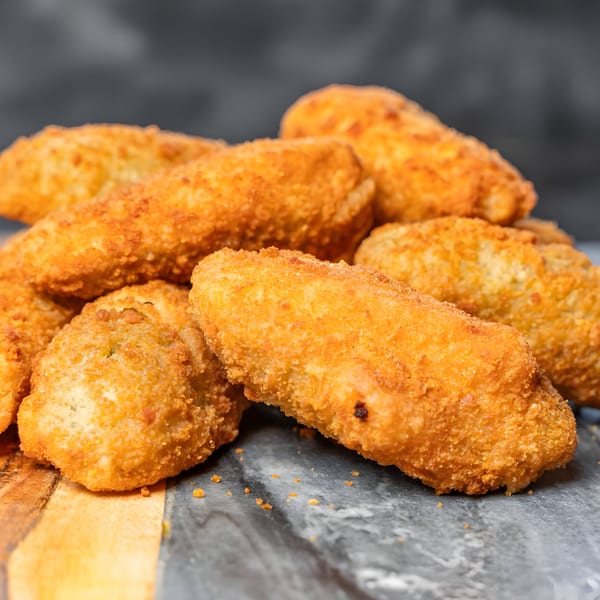 A close-up of golden-brown, breaded chicken tenders or croquettes piled on a dark, textured surface.