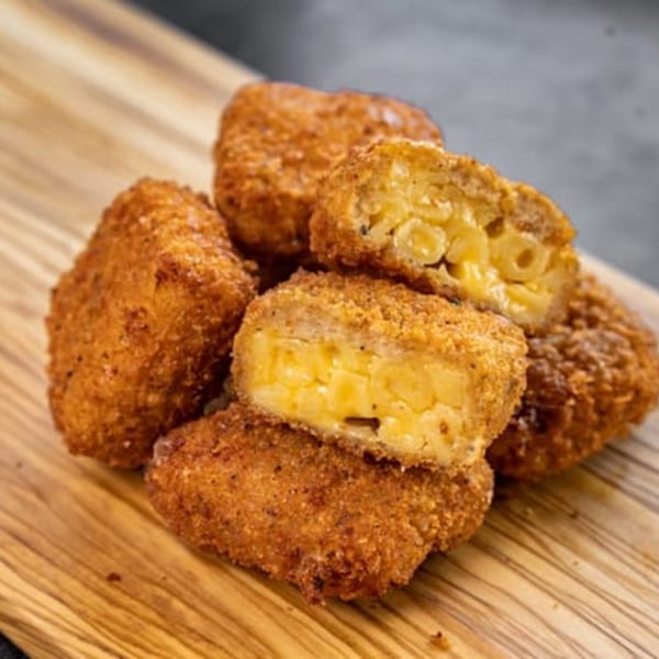 Golden-brown, breaded mac and cheese bites, one cut open to reveal creamy pasta inside, on a wooden board.