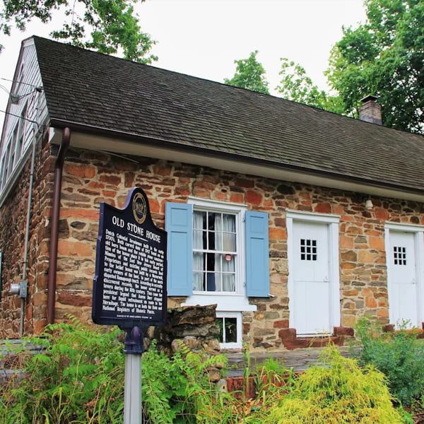 Historic stone house with a dark roof, light blue shutters, white doors, and a black sign.