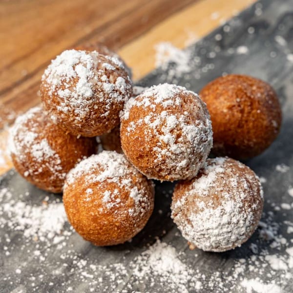 Six golden-brown donut holes, generously dusted with powdered sugar, on a dark surface.