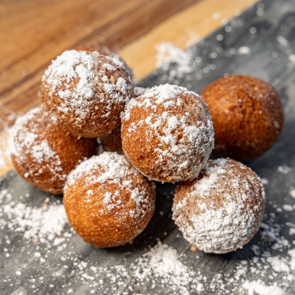 Six golden-brown donut holes, generously dusted with powdered sugar, on a dark surface.