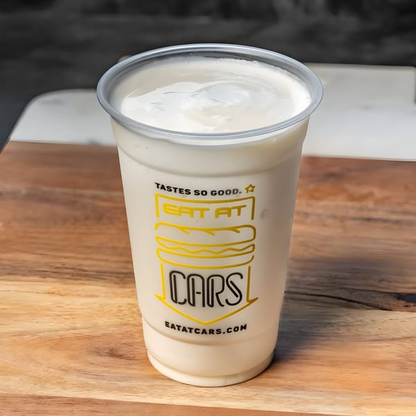 A vanilla milkshake in a clear plastic cup with the CARS Eatery logo on a wooden table.