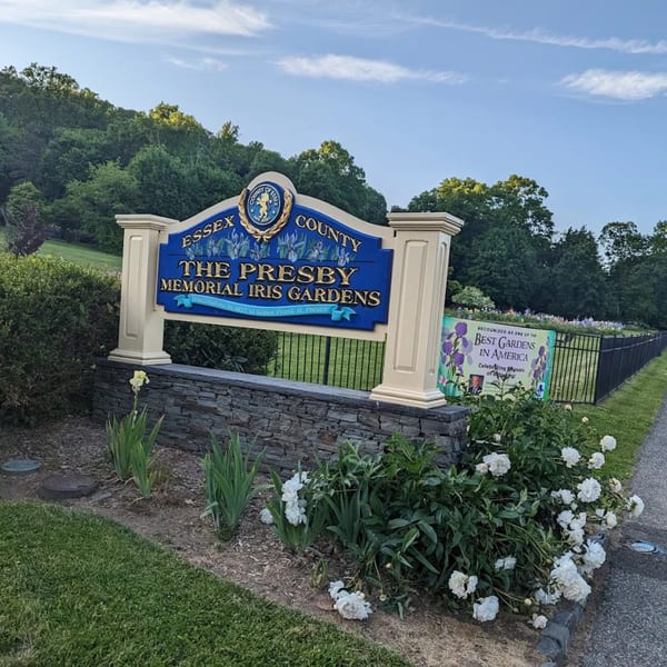A blue and gold sign for The Presby Memorial Iris Gardens, with white flowers and green foliage.